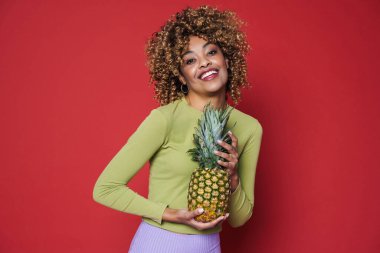 Young black woman laughing while posing with pineapple isolated over red background