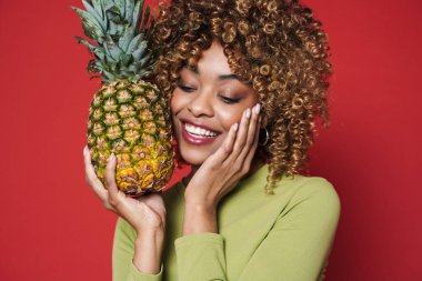 Young black woman laughing while posing with pineapple isolated over red background