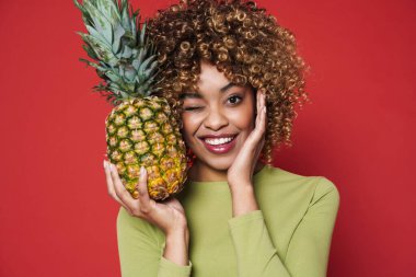 Young black woman winking while posing with pineapple isolated over red background