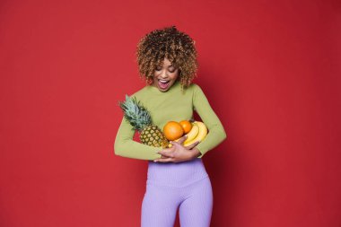 Young black woman expressing surprise while posing with fruits isolated over red background