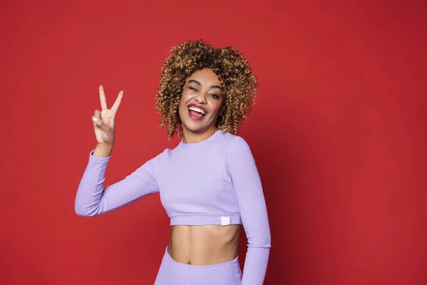 Young black woman with afro curls smiling and gesturing peace sign isolated over red background