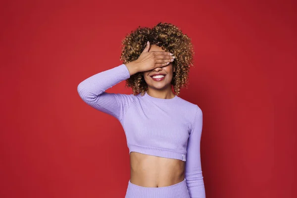 Young black woman with afro curls smiling at camera and covering her eyes isolated over red background