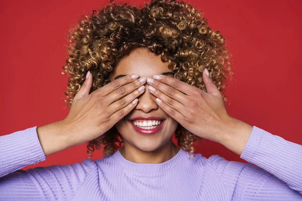 Young black woman with afro curls smiling at camera and covering her eyes isolated over red background