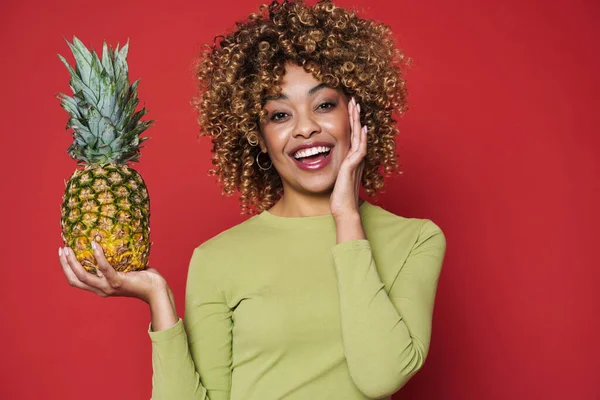 Young black woman laughing while posing with pineapple isolated over red background