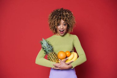 Young black woman expressing surprise while posing with fruits isolated over red background