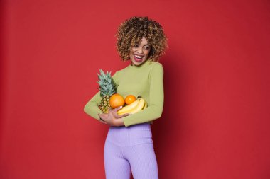 Young black woman laughing while posing with fruits isolated over red background