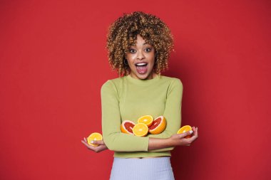 Young black woman with afro curls having fun holding oranges cut in half isolated over red background