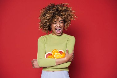 Young black woman with afro curls having fun holding oranges cut in half isolated over red background