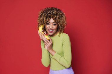 Young black woman laughing while posing with banana isolated over red background