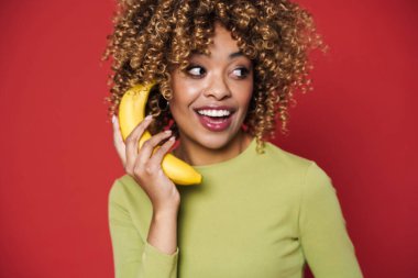 Young black woman laughing while posing with banana isolated over red background