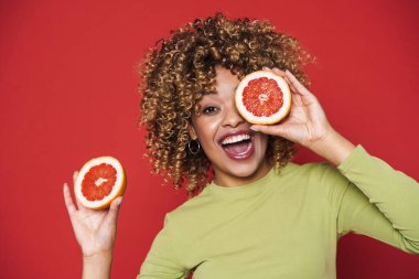 Young black woman with afro curls having fun holding orange cut in half isolated over red background