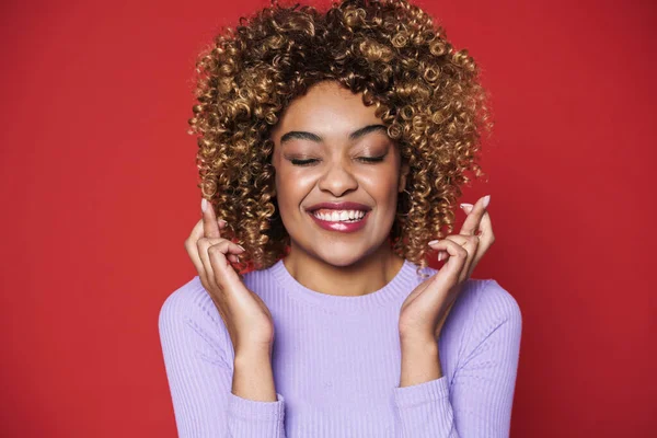 Young black woman with afro curls smiling and holding fingers crossed isolated over red background