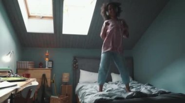 Funny curly haired young African girl listening music in headphones and dancing on the bed at home