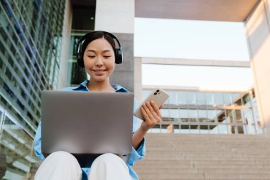 Asian woman in headphones using gadgets while sitting on stairs outdoors