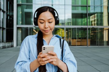 Asian brunette woman in headphones using cellphone while walking on city street