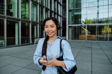 Asian brunette woman in headphones using cellphone while walking on city street
