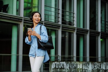 Asian brunette woman in headphones using cellphone while walking on city street