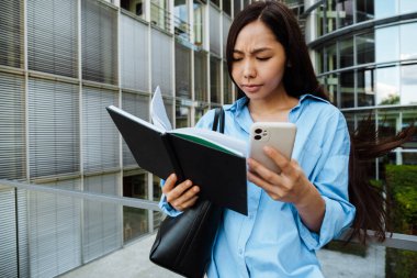Asian brunette woman using mobile phone while standing with planner outdoors