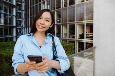 Asian brunette woman laughing and using mobile phone outdoors