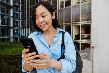 Asian brunette woman laughing and using mobile phone outdoors
