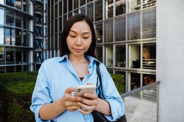 Asian brunette woman using mobile phone while standing by building outdoors