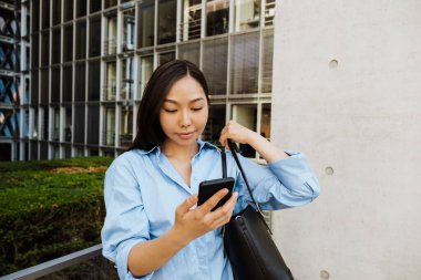 Asian brunette woman using mobile phone while standing by building outdoors