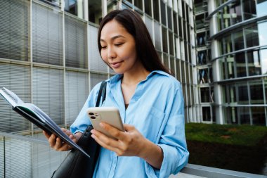 Asian brunette woman using mobile phone while standing with planner outdoors