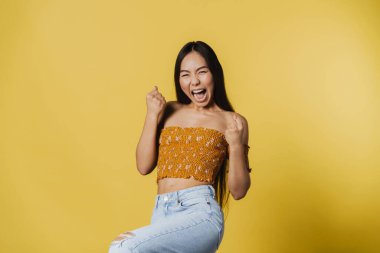Young asian woman screaming while making winner gesture isolated over yellow background