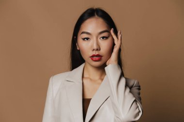 Young asian woman wearing jacket posing and looking at camera isolated over beige background