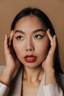 Young asian woman wearing jacket posing and looking at camera isolated over beige background