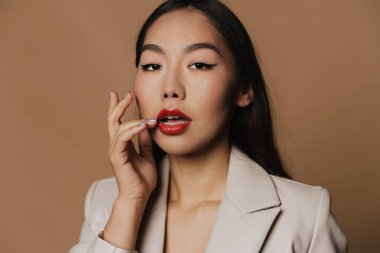 Young asian woman wearing jacket posing and looking at camera isolated over beige background