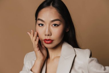 Young asian woman wearing jacket posing and looking at camera isolated over beige background