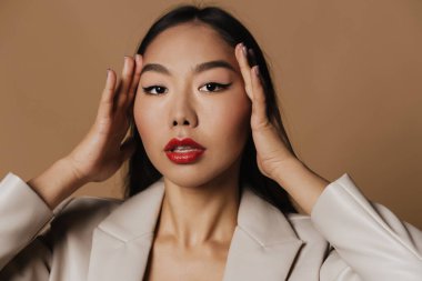 Young asian woman wearing jacket posing and looking at camera isolated over beige background