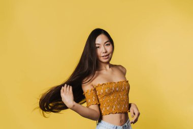 Young asian woman wearing top smiling while playing with her hair isolated over yellow background