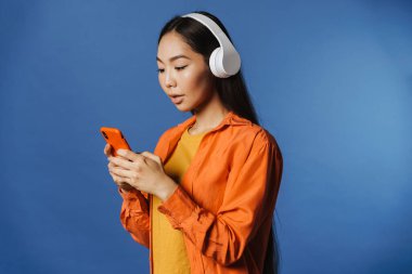 Young asian woman wearing headphones listening to music on cellphone isolated over blue background