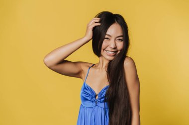 Young asian woman wearing dress smiling and looking at camera isolated over yellow background