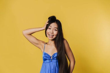 Young asian woman wearing dress smiling and looking at camera isolated over yellow background