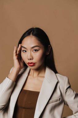 Young asian woman wearing jacket posing and looking at camera isolated over beige background