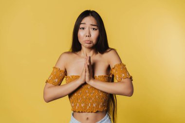 Young asian woman frowning while holding hands together isolated over yellow background