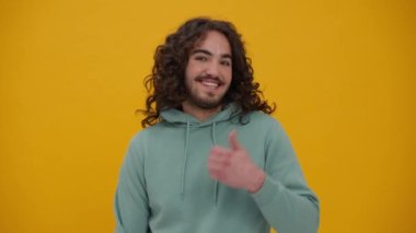 Smiling long-haired man in a blue hoodie talking at the camera and pointing to the copyspace in the yellow studio