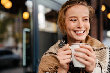 Joyful young blonde woman in warm plaid drinking hot tea while stanging outdoors at city street