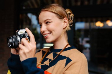 Cheerful blonde woman photographer in sweater taking photo on retro camera while walking outdoors