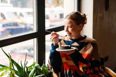 Joyful woman in colorful sweater eating delicious dessert while sitting at table in cafe