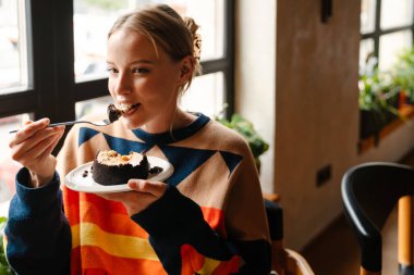Joyful woman in colorful sweater eating delicious dessert while sitting at table in cafe