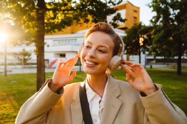 Portrait of young beautiful smiling happy woman in headphones looking aside, while standing on sunny lawn in the city