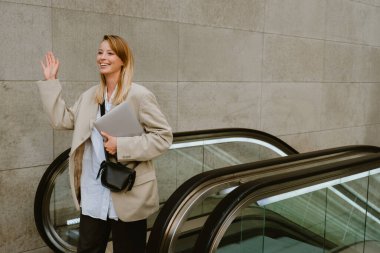Young beautiful smiling lady in blazer with laptop looking aside and waving , while getting off the escalator