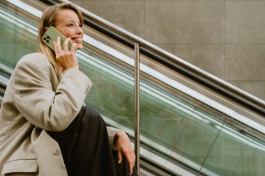 Young beautiful happy smiling business lady talking her phone and looking aside, while sitting on the stairs outdoors