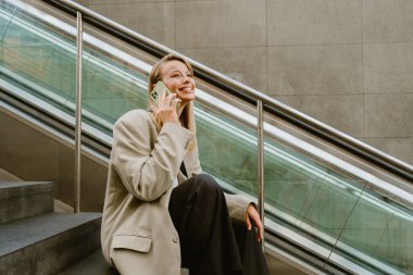 Young beautiful happy smiling business lady talking her phone and looking aside, while sitting on the stairs outdoors