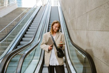 Young beautiful smiling business lady in blazer with laptop holding and using her phone, while standing near escalator