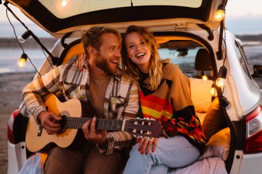 Happy young white couple smiling and playing guitar while sitting in trunk by seaside on sunset